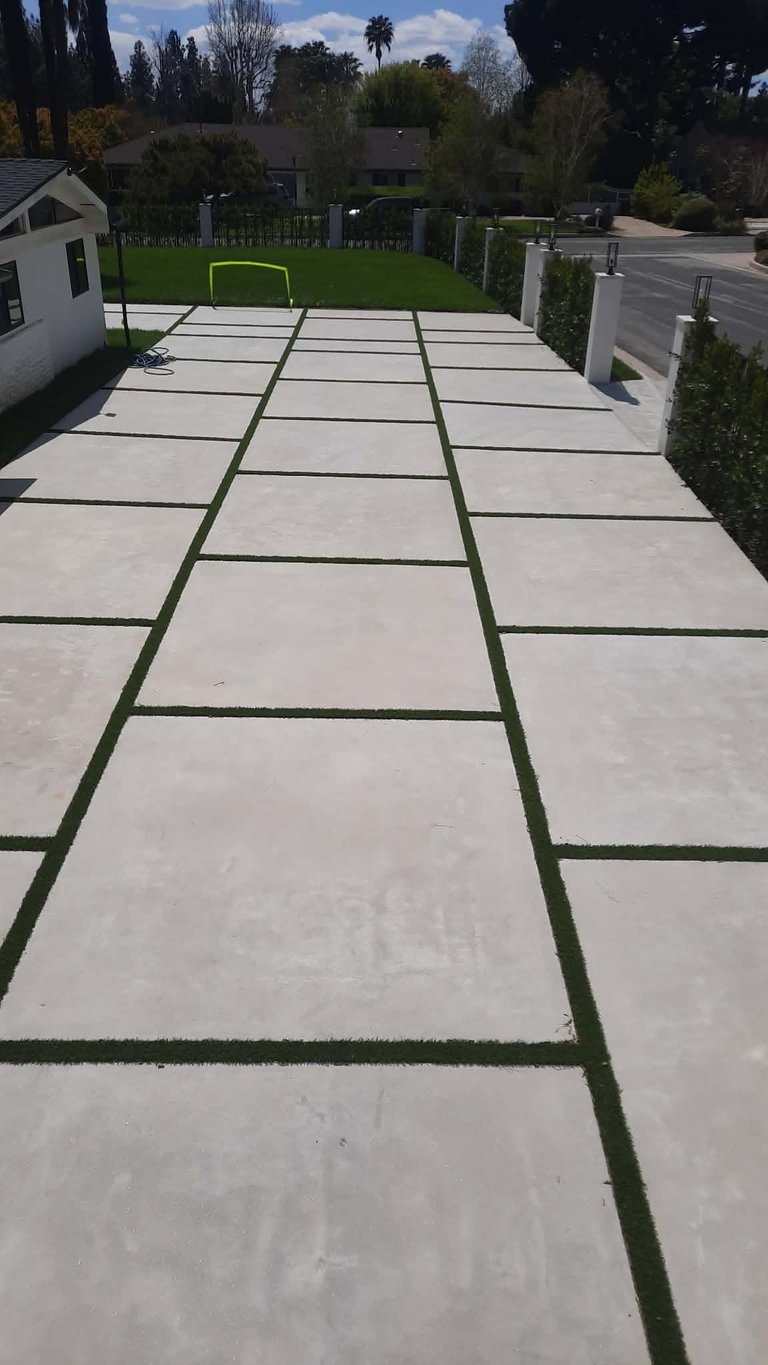 Modern concrete patio with green lawn strips between pavers, leading toward a residential backyard with white modern home and lush trees