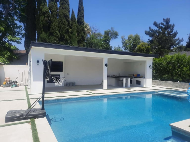 Modern white poolhouse next to a bright blue swimming pool with basketball hoop, surrounded by tall trees on a sunny day