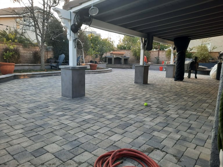 Paved outdoor courtyard with a modern pergola roof, concrete pillars, potted plants, and trees in background